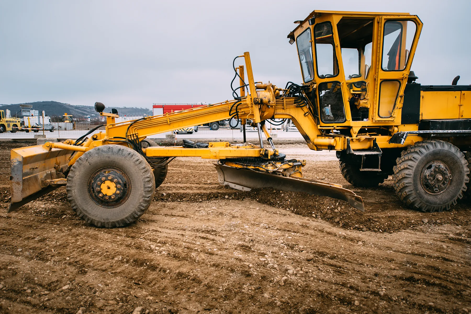 industry-details-construction-site-yellow-bulld-2024-09-16-21-43-32-utc Yellow motor grader leveling soil at a construction site, preparing the ground for infrastructure development under overcast skies.