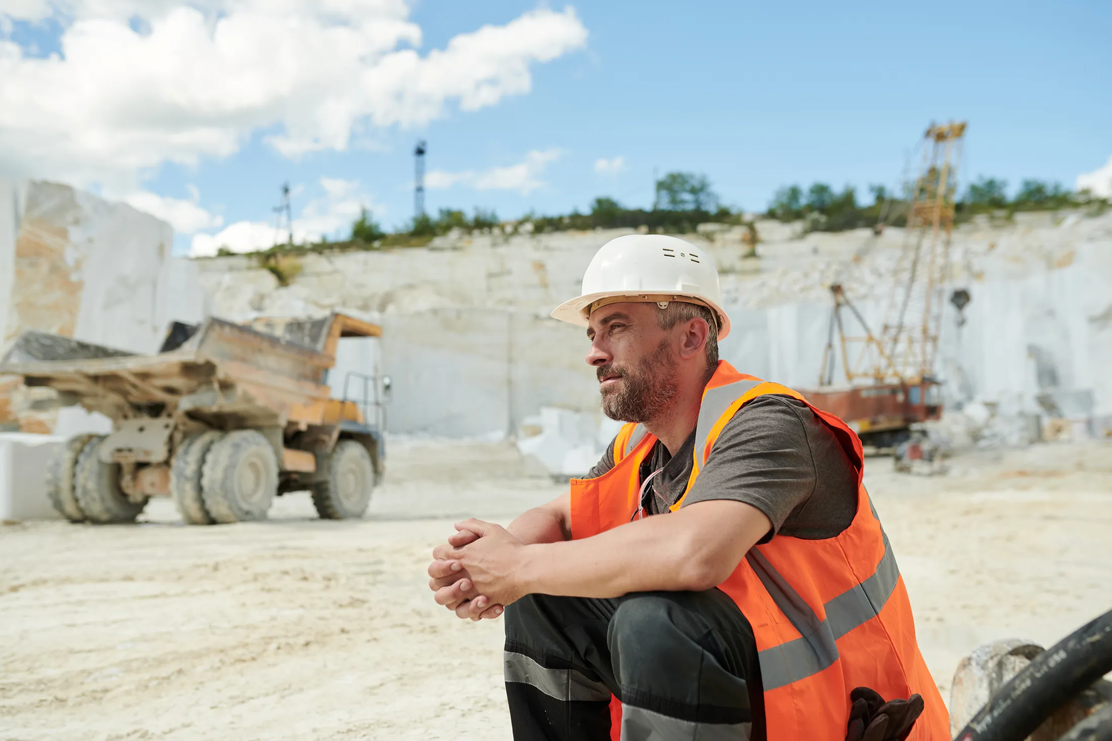 tired-worker-of-marble-quarry-sitting-in-front-of-2025-03-13-12-09-09-utc-1 Tired worker of marble quarry sitting in front of camera by workplace against dump truck and working bulldozer with crane