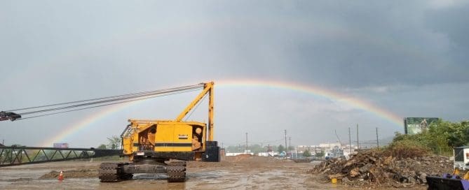 A yellow crawler crane on a muddy construction site with a double rainbow arching across a grey sky in the background.