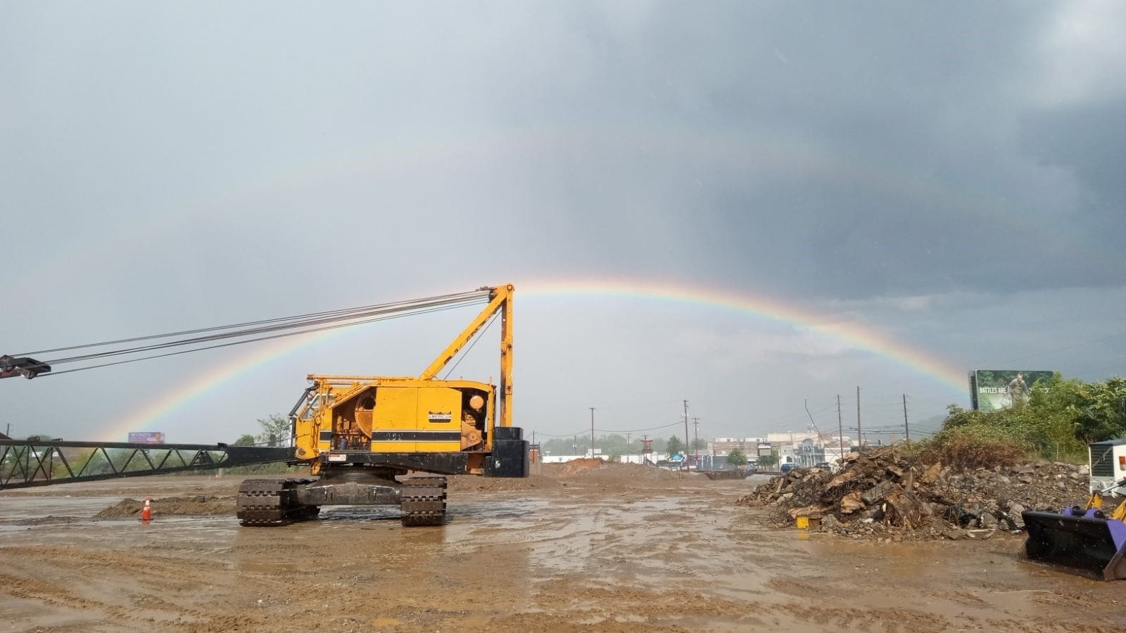 Yellow crawler crane on a muddy construction site with a vivid double rainbow arching over the background after a rainstorm.