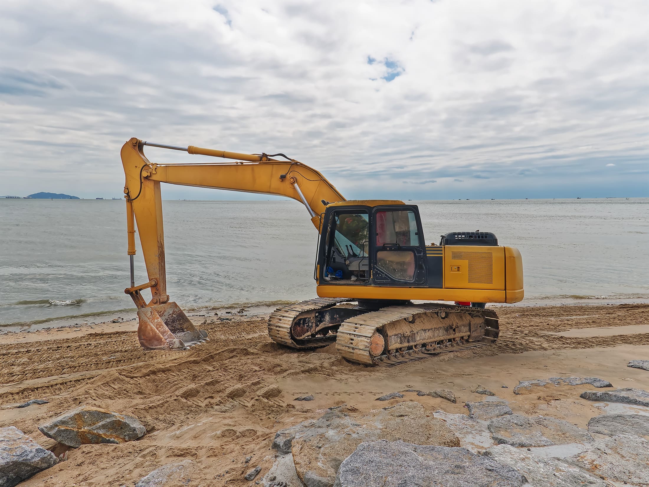yellow-excavator-working-on-the-beach-against-sea-2024-05-27-01-12-27-utc Yellow excavator parked on a sandy beach near the ocean, with tire tracks in the sand and overcast skies above.