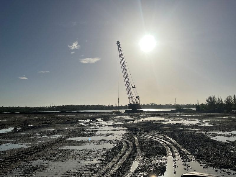 Crawler crane on muddy construction site at sunrise near a waterbody, with deep tire tracks and reflective puddles in the foreground.
