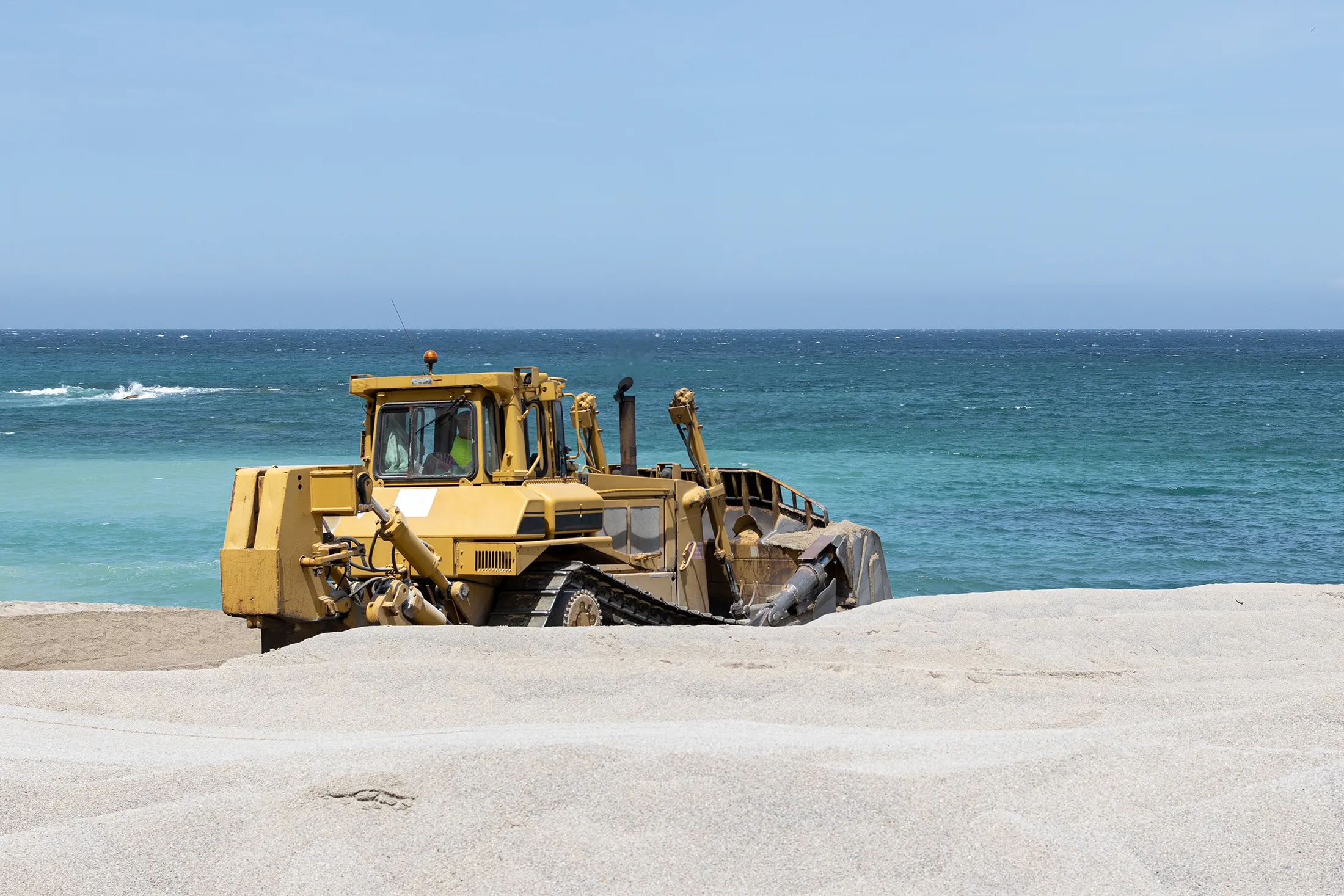 Bulldozer grading sand along a coastal construction site, illustrating soil stabilization and ground improvement work near shoreline structures.