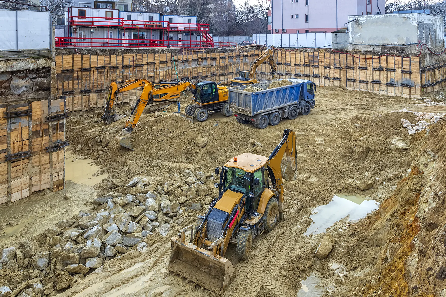 Excavators and trucks preparing a construction site near existing structures, illustrating soil excavation and ground improvement work in an urban environment.