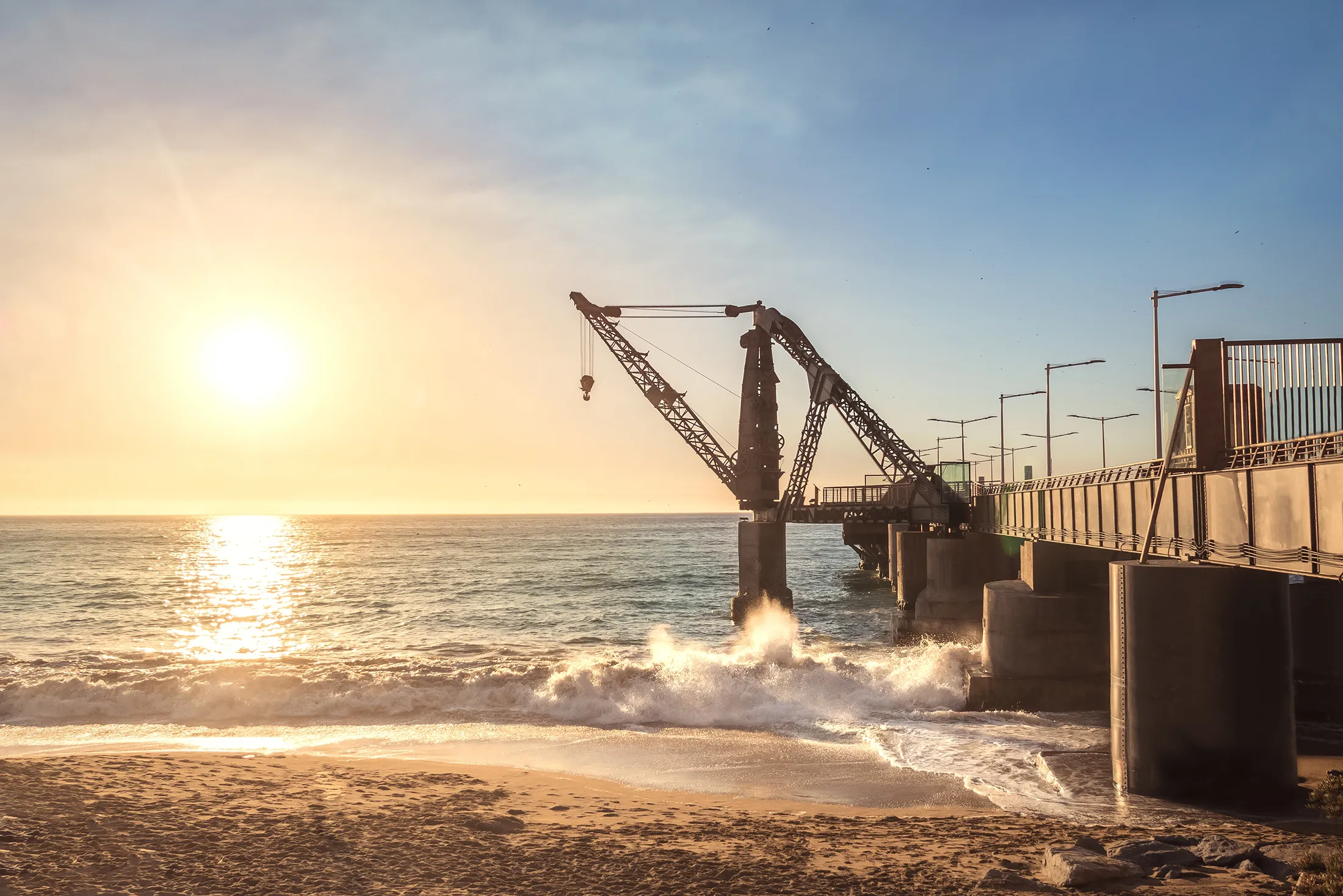 Sunset view of a coastal pier with heavy lifting cranes extending over the ocean, waves crashing against the structure along a sandy shoreline.