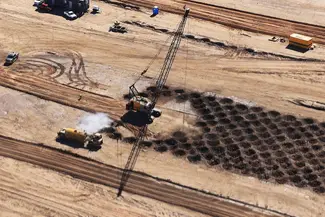 Aerial view of a crane performing dynamic compaction across a large grid pattern with visible impact craters on a construction site.